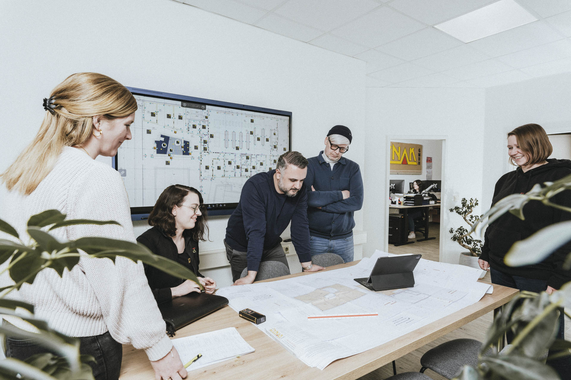 Teammeeting bei Oynak GmbH: Mitarbeitende besprechen gemeinsam Baupläne im Büro mit Bildschirm und Tablet – Fokus auf Teamarbeit und Projektplanung.